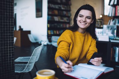 Elinde cep telefonu olan genç, pozitif esmer defter ile rahat bir kitap kafede latte içerken masada oturuyor.