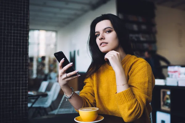 Charming young woman with dark hair in yellow sweater messaging on mobile phone while resting at table with cup of coffee