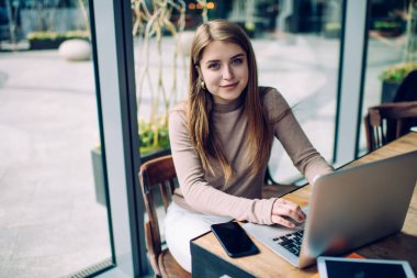 Yüksek açılı genç kız öğrenci, cam duvara karşı rahat kafeteryada otururken dizüstü bilgisayarla proje üzerinde çalışıyor.