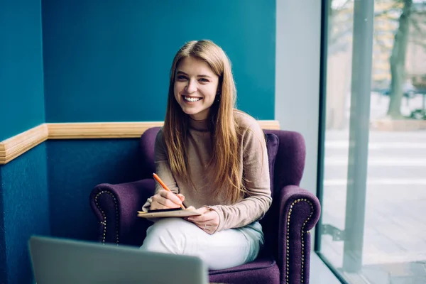 Young female student in casual outfit sitting in cozy soft armchair and writing notes on clipboard while looking at camera and smiling