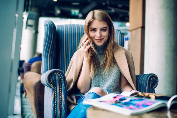 現代的なカフェで快適なアームチェアに座ってスマートフォンで話している間 流行の服を着て笑顔とカメラを見てのコンテンツの女性 — ストック写真
