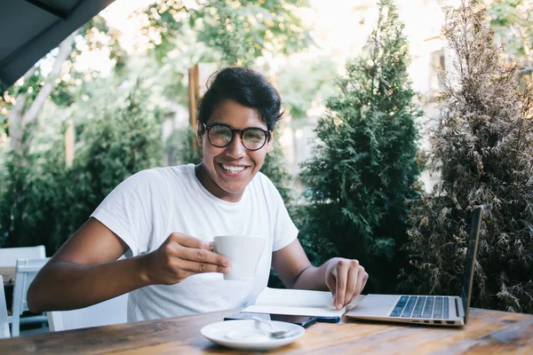 Cheerful male IT professional with cup smiling at camera sitting at street coffee shop with good internet connection