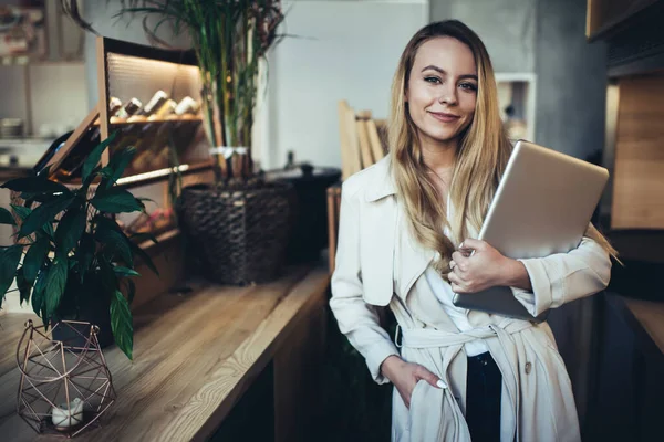 Happy young female entrepreneur in stylish coat with hand in pocket and laptop standing near table and looking at camera