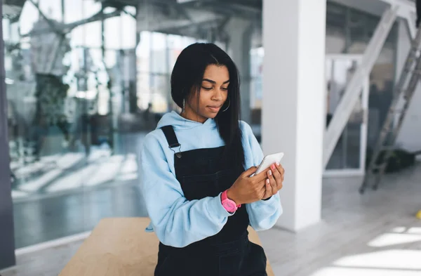 Black girl watching something on mobile phone while standing by wooden table. Concept of modern woman lifestyle. Young female person wearing casual clothes and wristwatch. Blurred interior of office