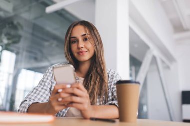 Masada cep telefonu kullanan Avrupalı bir kız. Modern başarılı kadın kavramı. Gündelik kıyafetler giyen ve kameraya bakan gülümseyen genç bir kadın. Defter, kalem ve kahve fincanı ahşap masada.