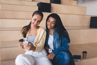 İş kadınları akıllı telefondan bir şeyler izler ve iş yerindeki ahşap banklarda kahve içerler. Avrupalı ve siyahi kızlar iş sırasında ara veriyorlar. Modern başarılı kadınlar. İş başında dinlenme fikri