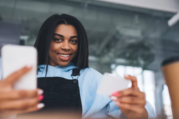 Smiling african girl holding business card and mobile phone. Concept of modern woman lifestyle. Selective focus of young female person wearing casual clothes. Lady looking at camera