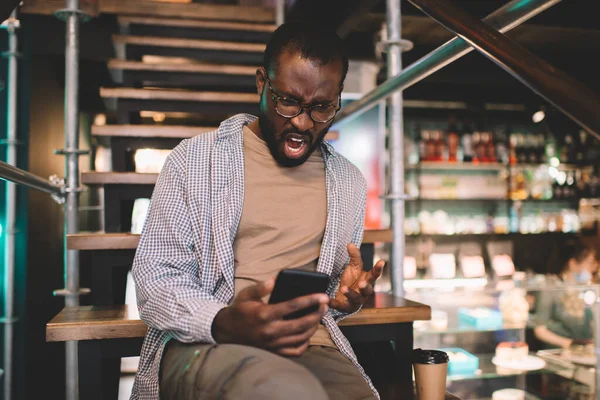 Unhappy dark skinned man in spectacles solving problems with internet connection on cellphone