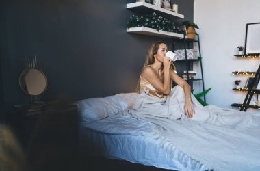 Avrupalı kız yatakta battaniyenin altında oturuyor ve evde fincandan çay ya da kahve içiyor. Ev hayatı kavramı. Dinlenme fikri. Sabahları pijamalı genç bir kadın. Modern yatak odasının içi.