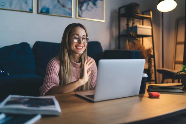 European girl waving hand during video call on laptop at home. Concept of domestic lifestyle. Idea of home rest and leisure. Young smiling woman in glasses sit at table. Interior of studio apartment