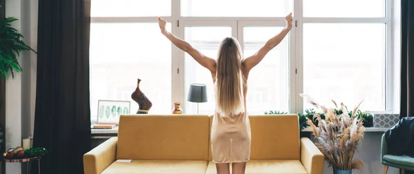 Back view of girl standing with open arms at home and looking at window. Concept of domestic lifestyle. Idea of rest. Woman in pajama waking up at morning time. Interior of bedroom in modern apartment
