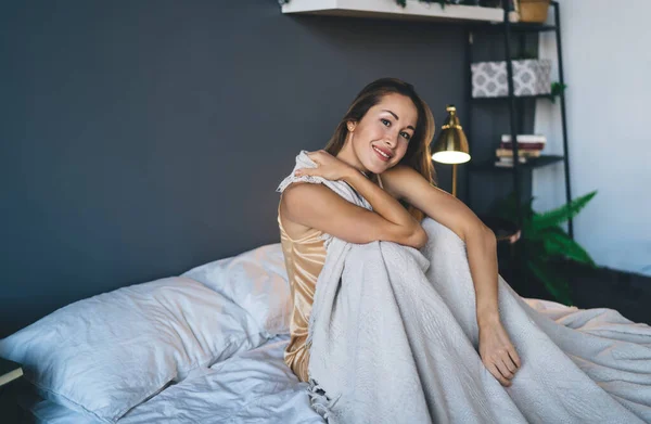 Smiling european girl sitting under blanket on bed and looking at camera at home. Concept of domestic lifestyle. Idea of rest. Young beautiful woman in pajama at morning time. Interior of bedroom
