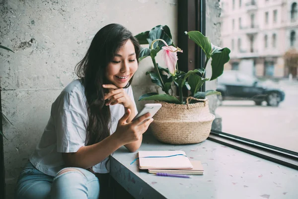 穿着蓝色牛仔裤 留着一头乌黑长发 满脸笑容的亚洲女士 白天坐在窗台上 带着仿真书 在现代智能手机上冲浪 — 图库照片