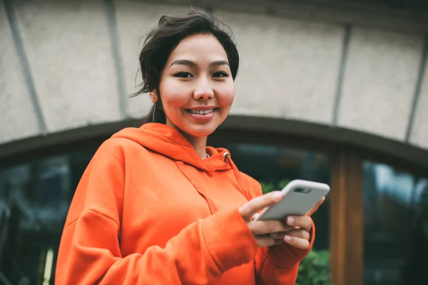 From below of happy Asian female in trendy hoodie text messaging on mobile phone while looking at camera with toothy smile