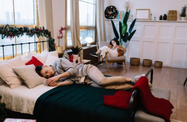 Genç çok ırklı çift evde dinleniyor ve akıllı telefon kullanıyor. Ev eğlencesi ve eğlence anlayışı. Asyalı kız koltukta oturuyor. Beyaz bir adam yatakta yatıyor. Geniş dairenin içi