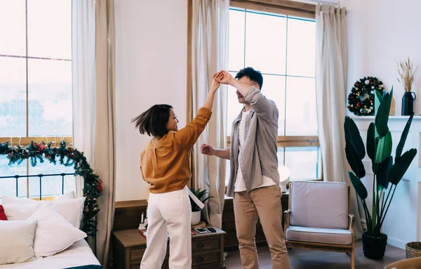 Young couple dancing at home during winter holidays. Romantic relationship and closeness. Man and smiling asian woman enjoying time together. Christmas and New Year time. Modern spacious apartment