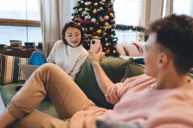 Adam cep telefonunda Asyalı kız arkadaşına dizüstü bilgisayarlı bir şey gösteriyor. Bir çift evdeki noel ağacının yanındaki kanepede dinleniyor. İlişki ve birlikte zaman geçirme kavramı. Noel ve Yeni Yıl Zamanı