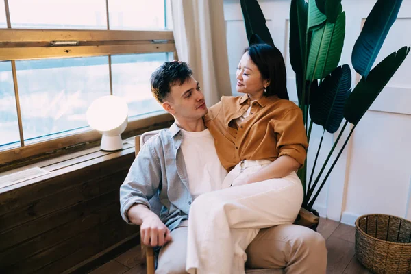 Asian woman sitting on knees of her boyfriend on armchair near window at home. Concept of romantic relationship and closeness. Multiracial couple enjoying time together and looking at each other