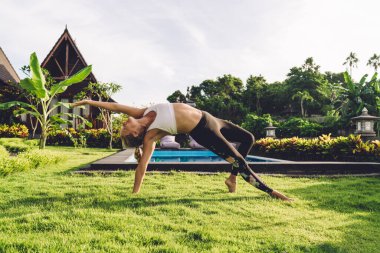 Spor kıyafetli sıska kadın, esneme hareketleri yapıyor ve yoga pozu veriyor. Kırsal evlerin yakınındaki yeşil çimlerde tek başına antrenman yapıyor.
