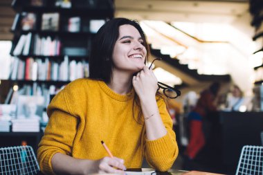 Sıcak kıyafetli gülümseyen kadın çalışanlar iş sırasında rahat bir kütüphanede otururken başka yere bakıyor ve başlangıç ödevini düşünüyorlar.