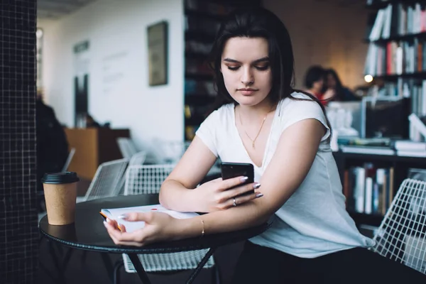 Συγκεντρωμένη Φοιτήτρια Περιήγηση Smartphone Και Κάθεται Στο Γραφείο Copybook Ενώ — Φωτογραφία Αρχείου