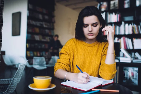 Concentrated young woman in casual clothes writing in planner while sitting at table in modern workplace with cup of hot coffee and creating plan