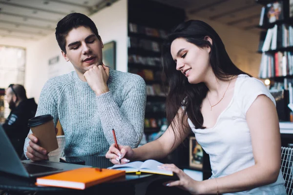 Young smart guy in casual clothes helping female friend with homework assignment while studying together in contemporary library with books and laptop