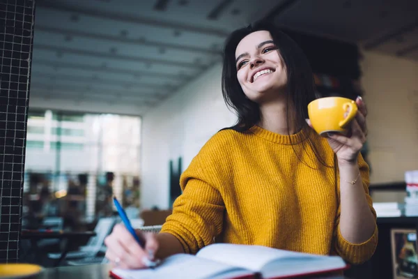 Low angle of positive young woman drinking coffee and writing notes in notebook while sitting alone at table in cafe