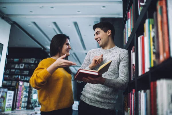 下面是一对微笑的夫妇在商店里交谈的情景 男人们手握打开的书的内容靠在书架上 女人则在书架上展示 — 图库照片