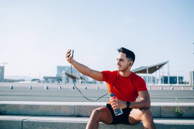 Kırmızı tişörtlü, mavi şortlu, neşeli, kaslı ve atletik bir erkek. Bir şişe suyla güneşli bir günde selfie çekiyor.