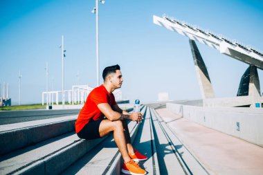 Kırmızı tişörtlü, mavi şortlu, granit merdivenlerde bir şişe suyla oturan modern kaslı atletik bir erkeğin yan görüntüsü.