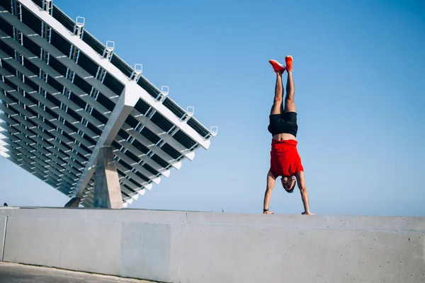 Energetic young male in red t shirt blue sportive shorts orange sneakers standing on hands on granite podium in stadium in summer