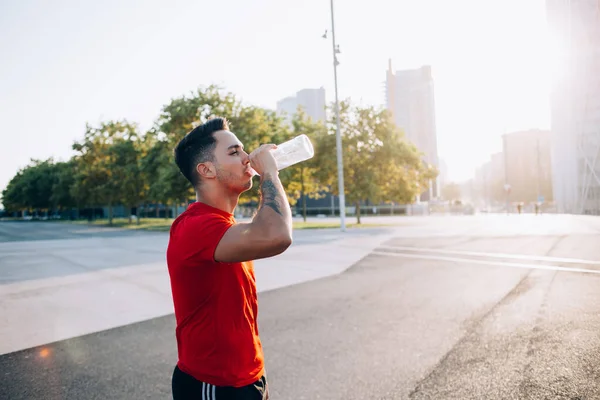 Thirsty athletic man holding bottle for cold refreshing after energetic outdoors jogging around urban setting