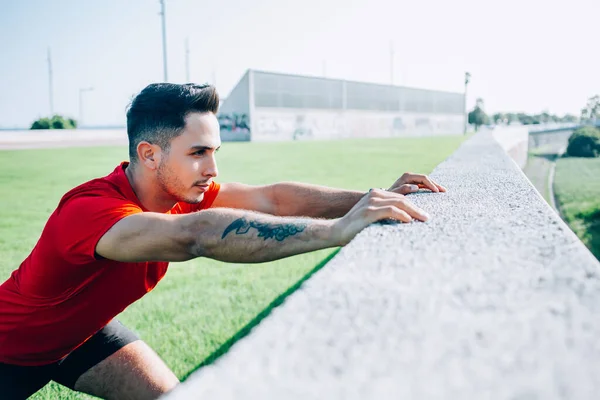 Concentrated ethnic young man in sportswear with tattoo preparing for jogging exercise stretching legs leaning hands on stone wall in stadium