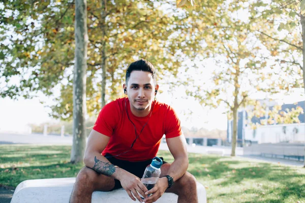 Muscular trainer man holding water bottle in hands listening audio records via electronic headphones during workout break