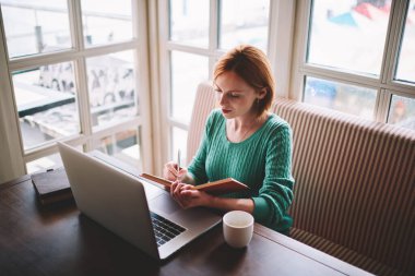 Gündelik kazaklı, modern dizüstü bilgisayarlı ve not defteriyle proje üzerinde çalışan genç ve ciddi bir kadının yüksek açısı.