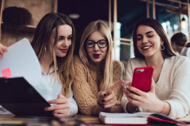 Gündelik kıyafetlerle gezen, modern bir kafede oturan ve akıllı telefonları karıştıran bir grup neşeli kadın.