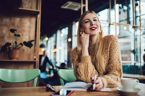 Cheerful female freelancer in casual outfit and red lipstick sitting at wooden table and looking at camera while taking notes in copybook