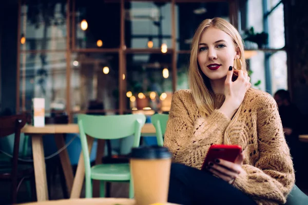 穿着保暖衣服的乐观的年轻女性坐在咖啡桌旁 一边喝着外卖咖啡 一边用智能手机把头发调整到全景窗口 — 图库照片