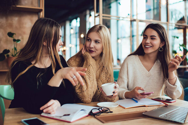 Оптимистичные молодые женщины улыбаются и смотрят друг на друга, сидя за столом с ноутбуком и ноутбуком и обсуждая новый проект во время встречи