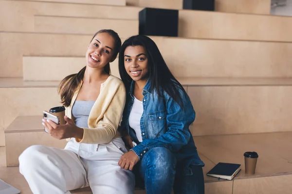 Businesswomen having break during work on wooden benches in coworking office space. Smiling european and black girls with coffee looking at camera. Modern successful women. Idea of resting on job