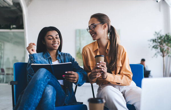 Multiracial businesswomen watching something in notebook. Concept of modern successful women. Idea of teamwork. Young smiling european and black girls sitting in chairs in office