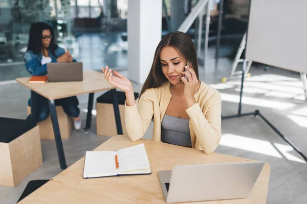 European businesswoman talking on mobile phone while her european black female colleague working on blurred background. Concept of modern successful women. Young girls sitting at desks in sunny office