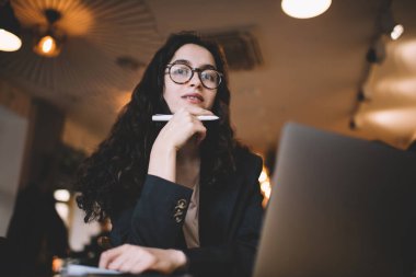 Masada dizüstü bilgisayarla otururken elinde kalem tutan, uzun kıvırcık saçlı, çekici kadın serbest yazardan.