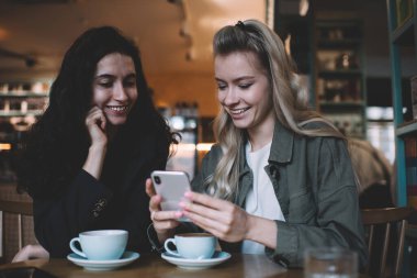 Kafede kahve fincanları ve cep telefonu kullanarak gündelik kıyafetlerini giyen pozitif çok ırklı kadın arkadaşlar.