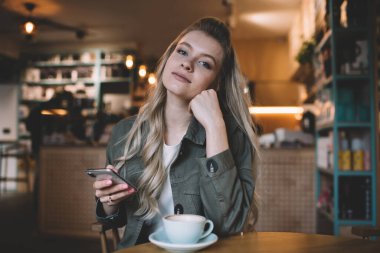 Dalgalı saçlı, gündelik elbiseli, genç, çekici bir kadın, ahşap masada, bir fincan sıcak kahve ve cep telefonuyla oturuyor.