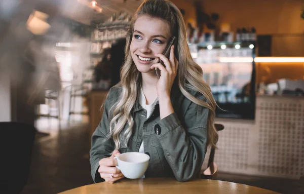 笑容可亲的年轻女士 留着波浪般的头发 在智能手机上说话 喜欢在模糊的背景下在现代咖啡馆里聊天和喝酒 — 图库照片