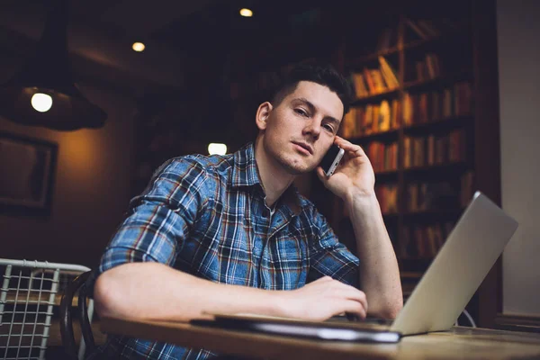 Portrait of male student in smart casual shirt using laptop device for doing distance e learning and smartphone gadget for making consultancy conversation