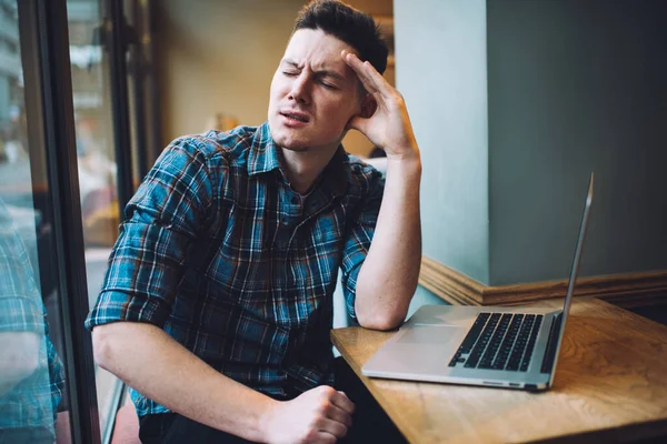 Unhappy male software developer disappointed with program code mistake sitting at table near window and feeling headache