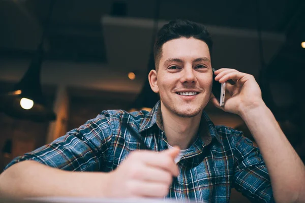 Portrait of cheerful hipster guy smiling at camera while calling to best friend for discussing meeting time in coffee shop